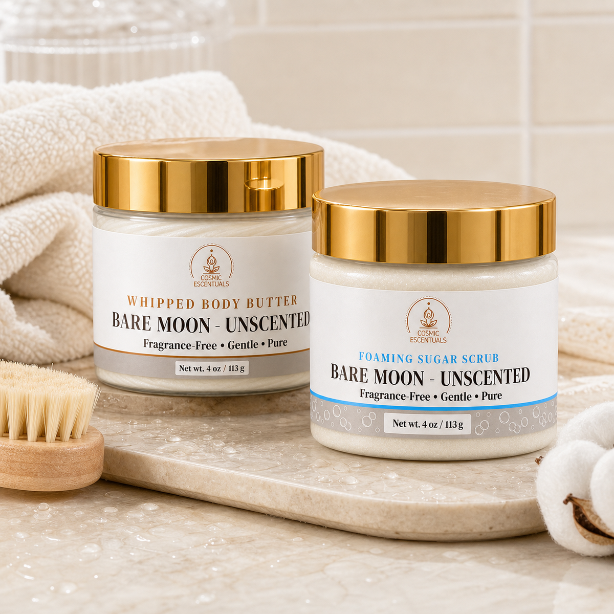Bare Moon Unscented Whipped Body Butter and Foaming Sugar Scrub jars with gold lids on a marble ledge, styled with a wooden body brush, a folded cream towel and cotton bolls.