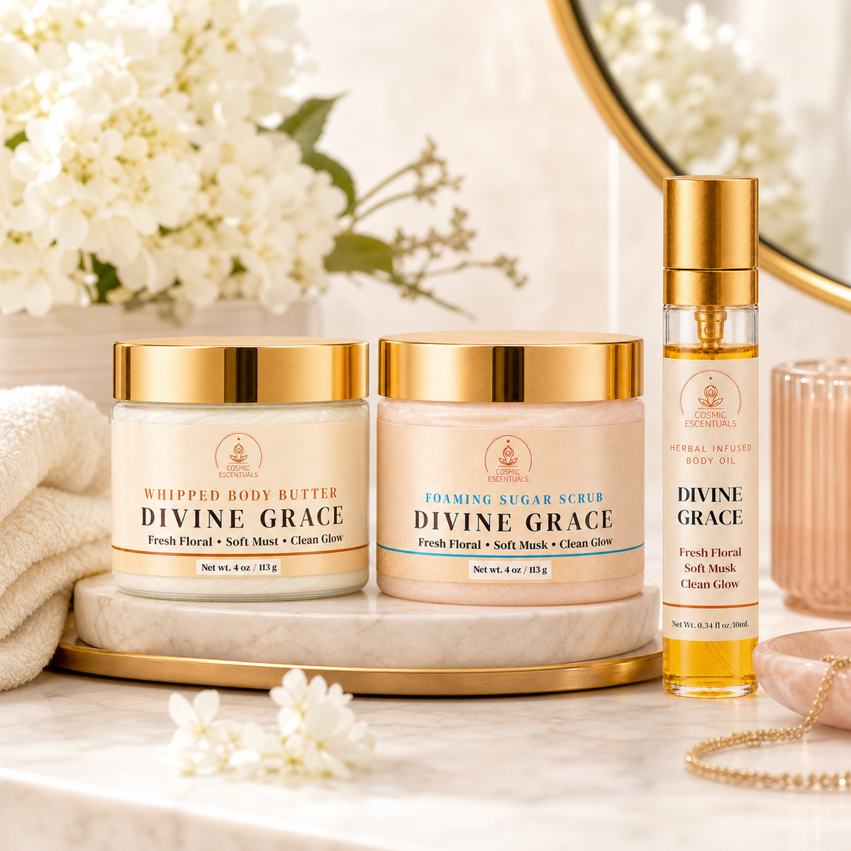 Three Divine Grace skincare products from Cosmic Escentuals are arranged on a marble tray with white flowers and a gold mirror in a bright, spa-like setting. The products include Whipped Body Butter, Foaming Sugar Scrub, and a Divine Grace Body Oil spray bottle.