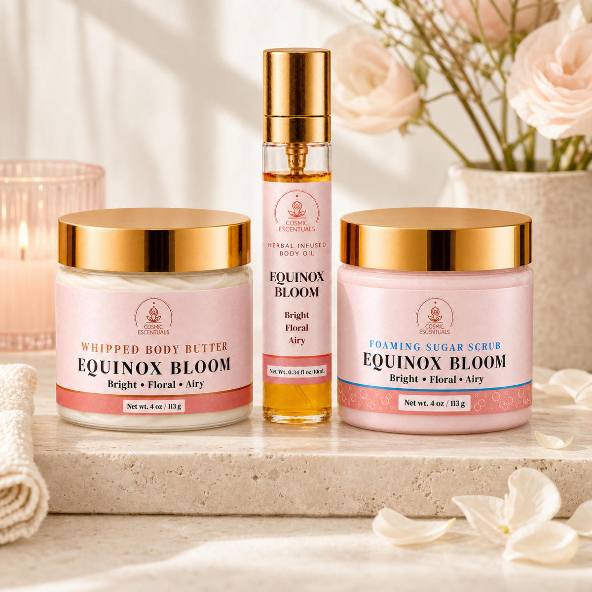 Equinox Bloom Whipped Body Butter, Herbal Infused Body Oil and Foaming Sugar Scrub in blush pink with gold lids, on a stone ledge with roses, a lit candle and a rolled towel.