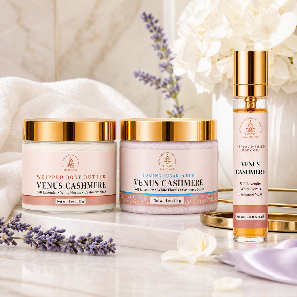 Three Venus Cashmere body care products arranged on a white marble surface with soft floral props: two jars labeled Whipped Body Butter and Foaming Sugar Scrub, plus a small body oil bottle. The products have pale pink labels with gold caps and gold lids, photographed in a bright, clean setting with white flowers and lavender sprigs.