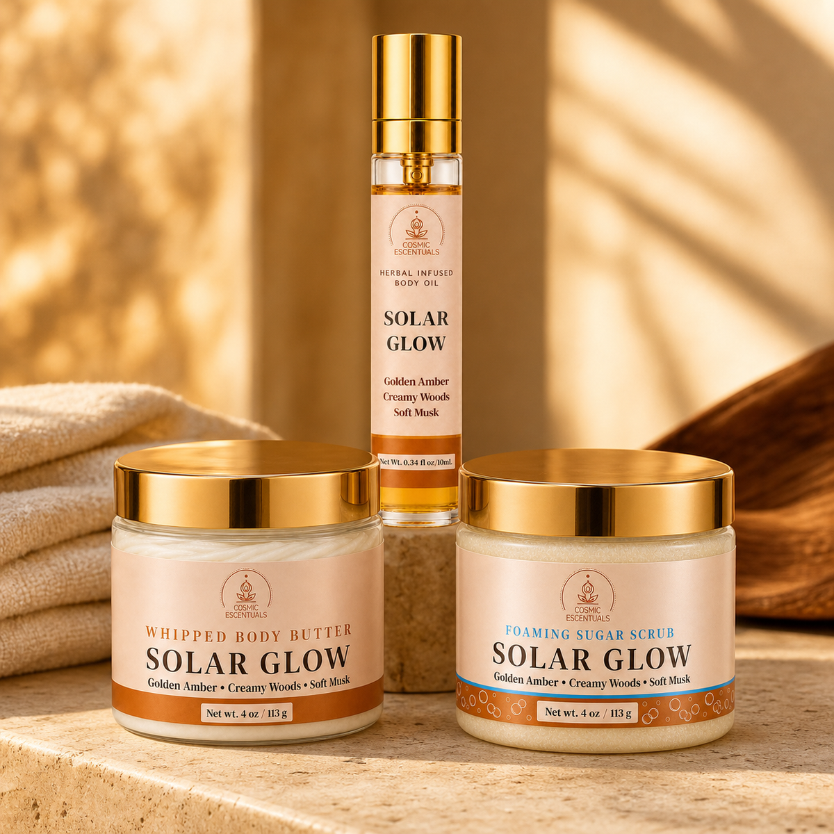 Three natural body care products from Cosmic Escentuals arranged on a stone surface: a slim spray bottle of Solar Glow body oil in the back, a jar of whipped body butter in the front left, and a jar of foaming sugar scrub in the front right. The products have cream-colored labels with gold lids and gold text, set against a warm, sunlit background with soft shadows.