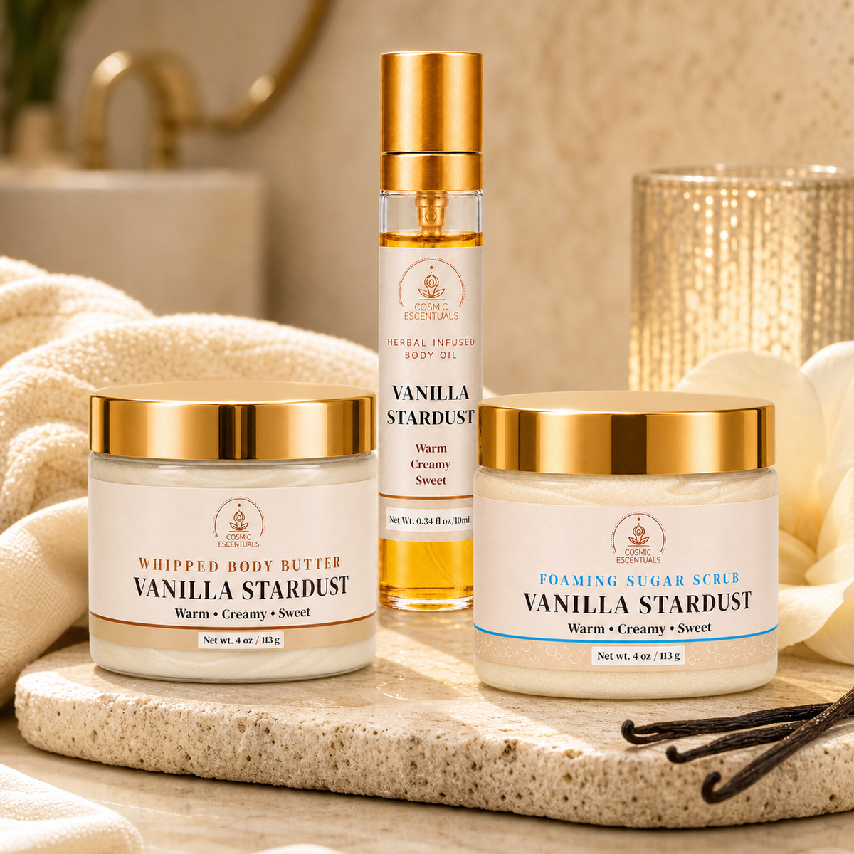 Three skincare products from Cosmic Escentuals are arranged on a stone tray in a warm bathroom setting: a whipped body butter jar, a foaming sugar scrub jar, and a body oil spray bottle. The products feature white labels with gold lids and gold accents, with vanilla beans and a textured gold vase in the background.