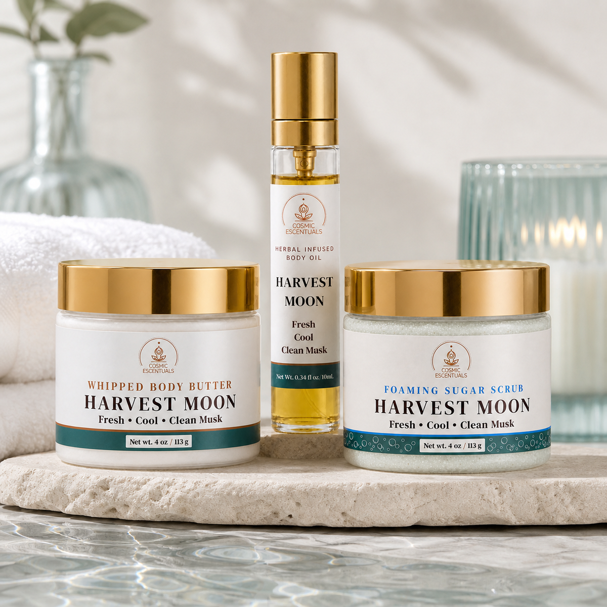 Three skincare products from Cosmic Escentuals under the Harvest Moon line are arranged on a stone tray: a whipped body butter jar, a body oil spray bottle, and a foaming sugar scrub jar. The products have white labels with gold lids and teal accents, set against a soft-focus background with a candle and glass.