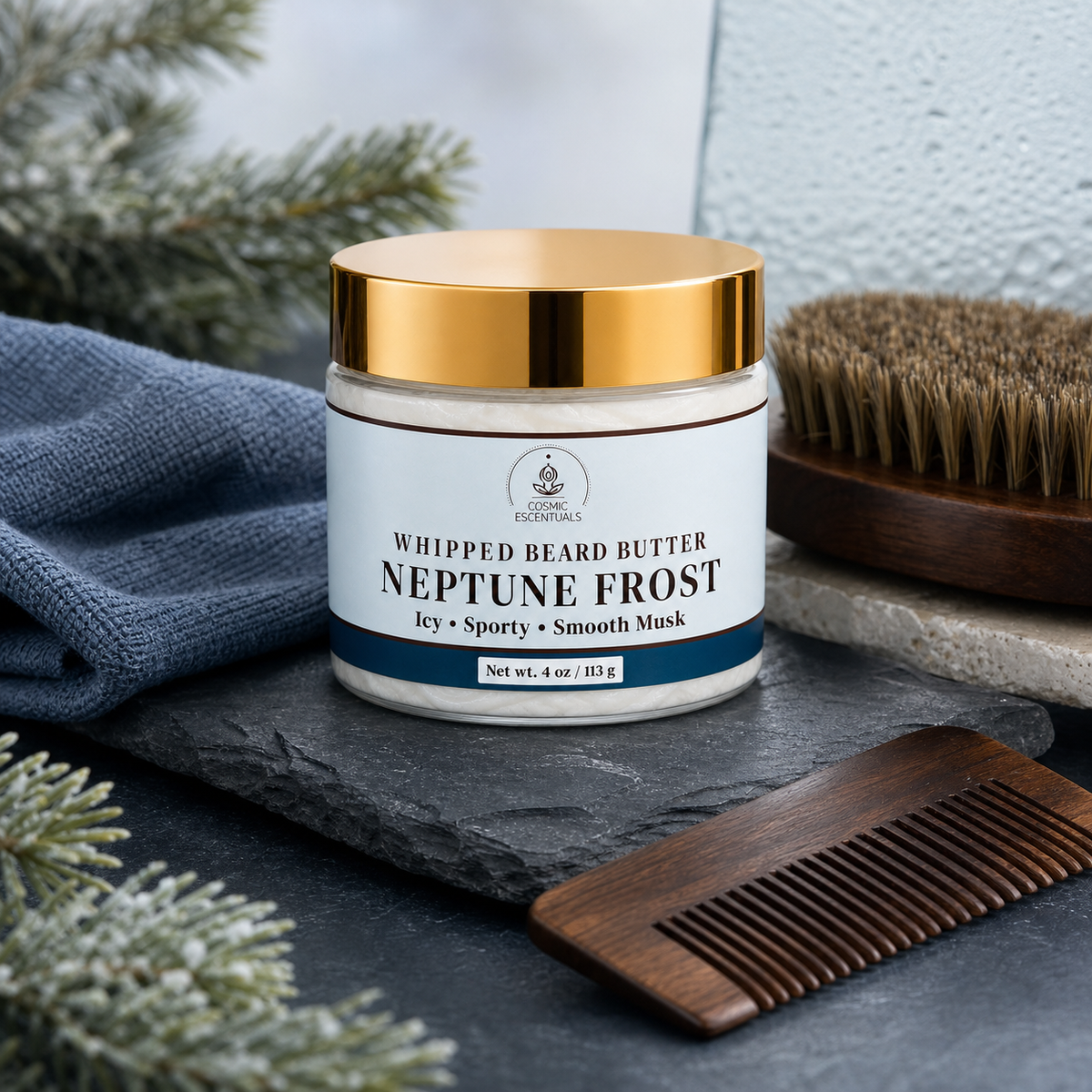 A jar of Whipped Beard Butter labeled Neptune Frost is styled on a dark slate surface with a wooden comb, a blue cloth, evergreen branches, and a beard brush in the background.