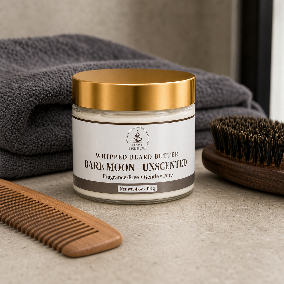A jar of Bare Moon Unscented Whipped Beard Butter by Cosmic Essentials, styled on a stone surface with a wooden comb, a dark gray towel, and a beard brush.