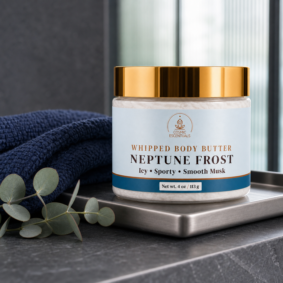 A jar of Whipped Body Butter in the Neptune Frost scent sits on a silver tray next to a navy blue towel and eucalyptus leaves. The jar has a white label with gold and blue accents and a shiny gold lid.