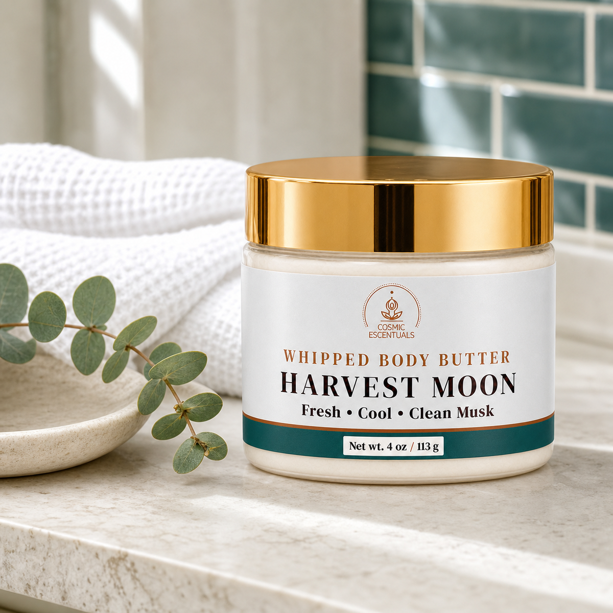 A jar of Organic Essentials Whipped Body Butter labeled 'Harvest Moon' sits on a marble countertop with a white towel, a small dish, and eucalyptus leaves in a bathroom setting.