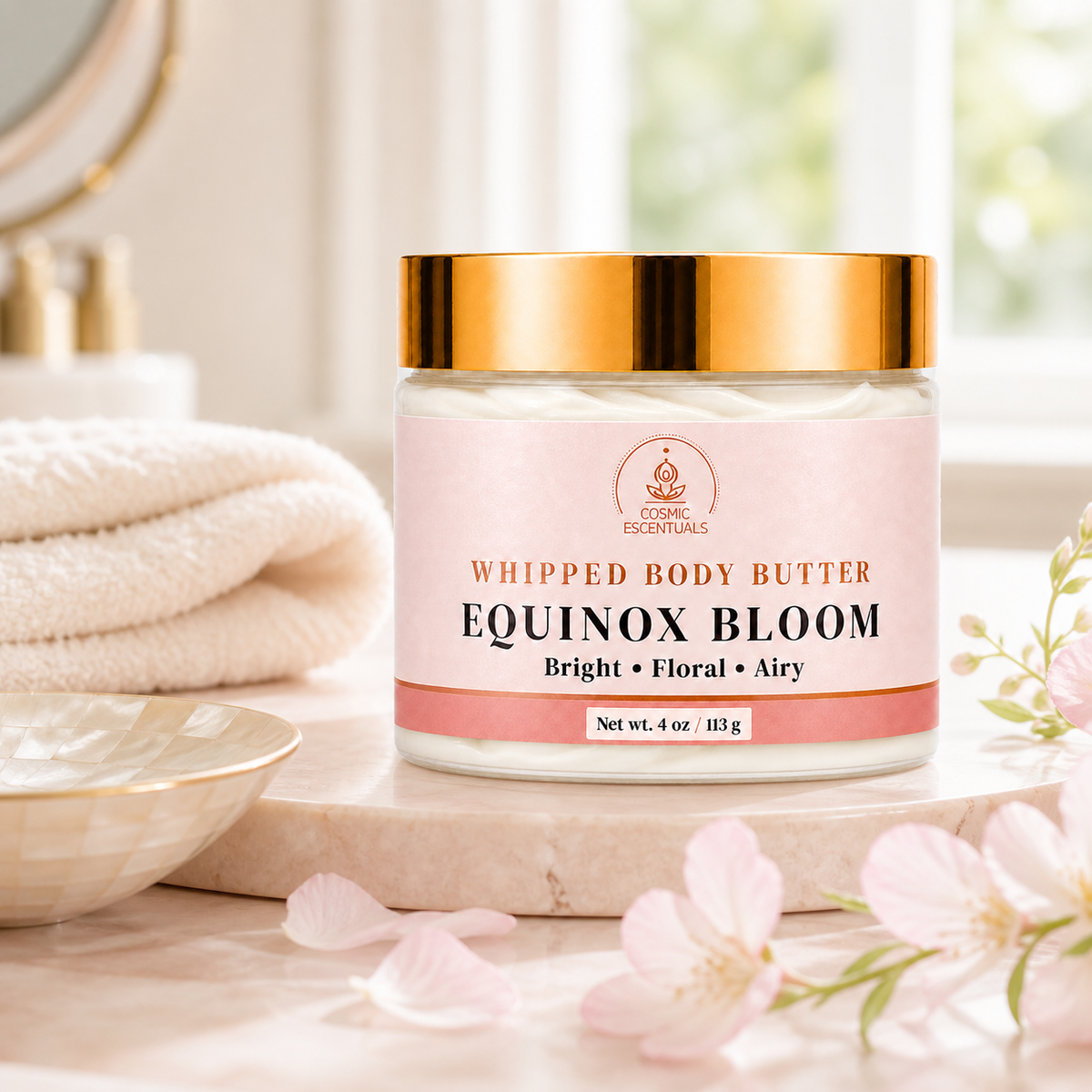 A jar of Cosmic Escentuals Whipped Body Butter in the Equinox Bloom scent, styled on a marble tray with folded towels, a bowl, and pink blossoms.