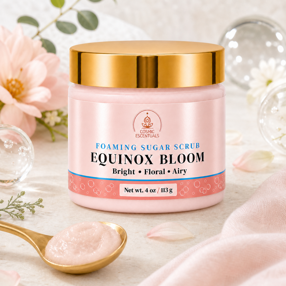 A jar of Cosmic Escentuals Equinox Bloom Foaming Sugar Scrub, featuring a pink body with a shiny gold lid, styled on a light beige fabric with a gold spoon holding some of the pink scrub, and soft pink flowers in the background.