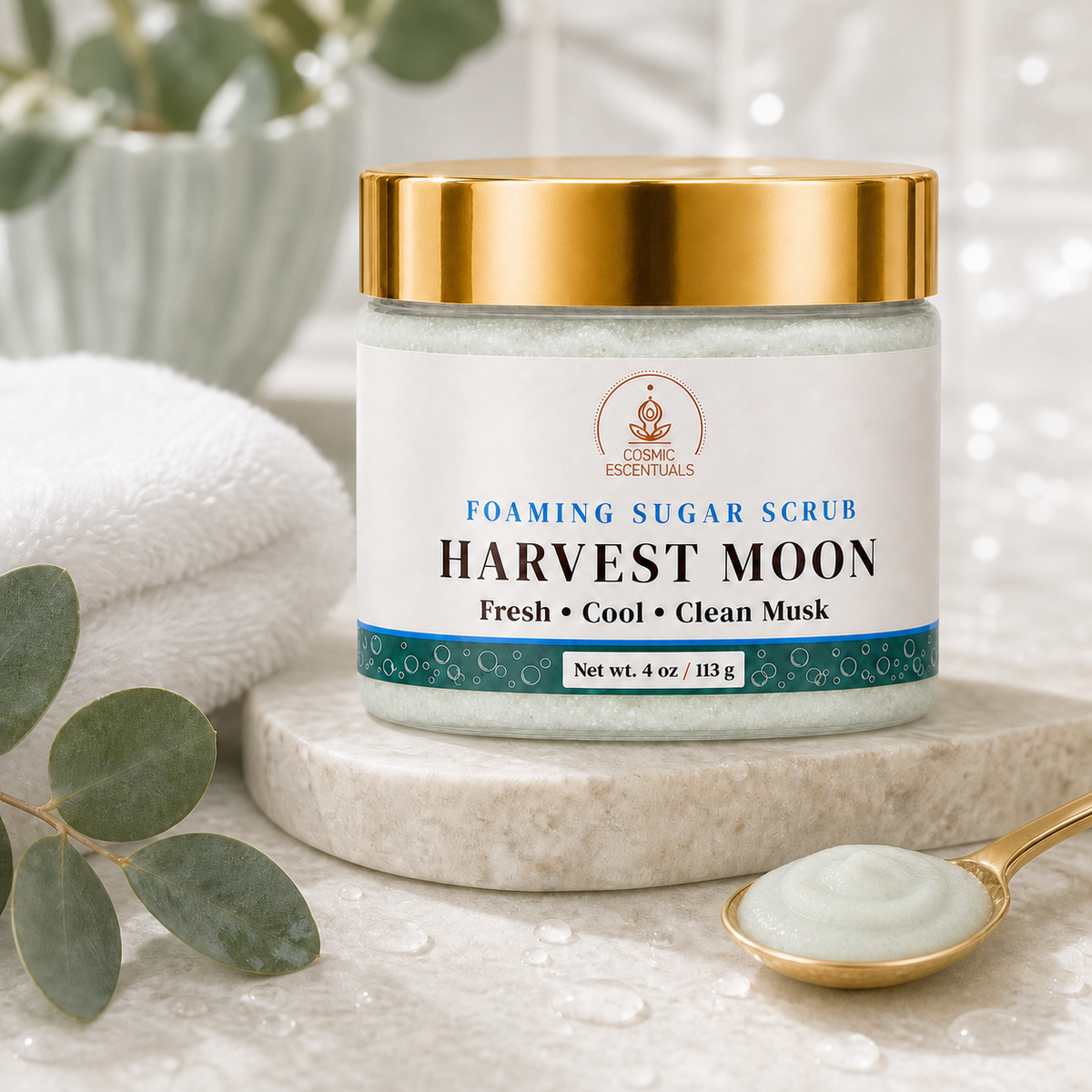 A jar of Chakra Essentials Harvest Moon Foaming Sugar Scrub sits on a marble pedestal with a gold spoon of scrub, white towels, and eucalyptus leaves in a bright, clean setting.