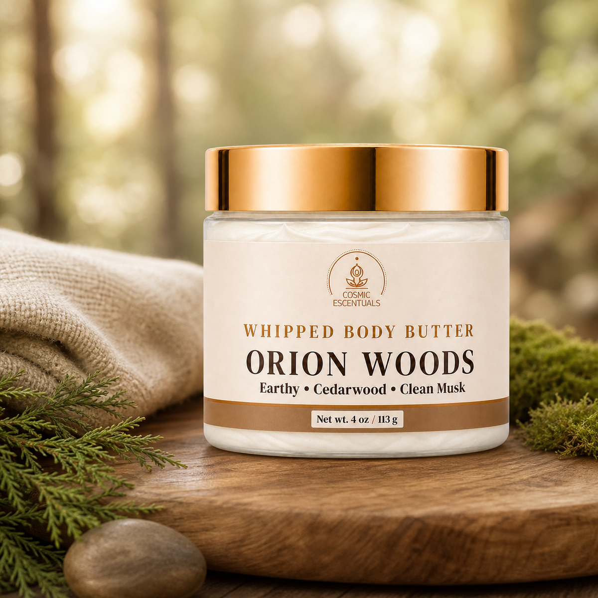 A jar of Organic Essentials Whipped Body Butter in the Orion Woods scent, styled on a rustic wooden surface with a linen cloth, pine needles, and moss against a soft-focus forest background.