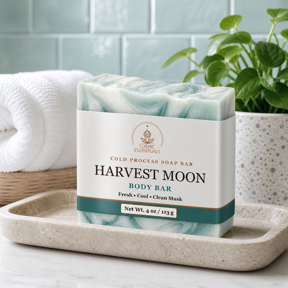 Harvest Moon Body Bar, swirled teal and white, on a stone dish with a rolled white towel and a potted plant against pale green tile.