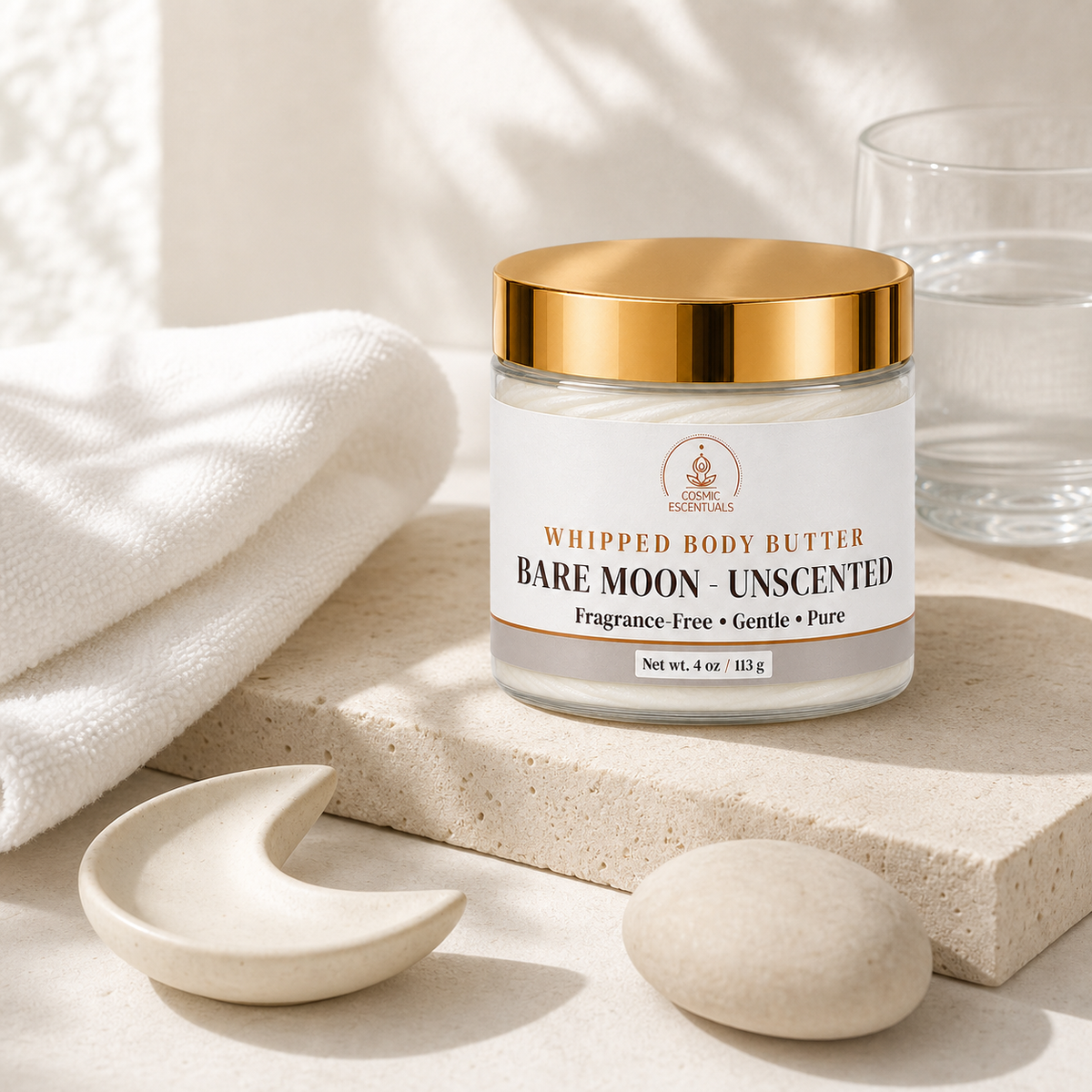 A jar of Bare Moon Unscented Whipped Body Butter by The Scented Shop, styled on a beige stone slab with a white towel, a small ceramic crescent dish, a stone, and a glass of water.