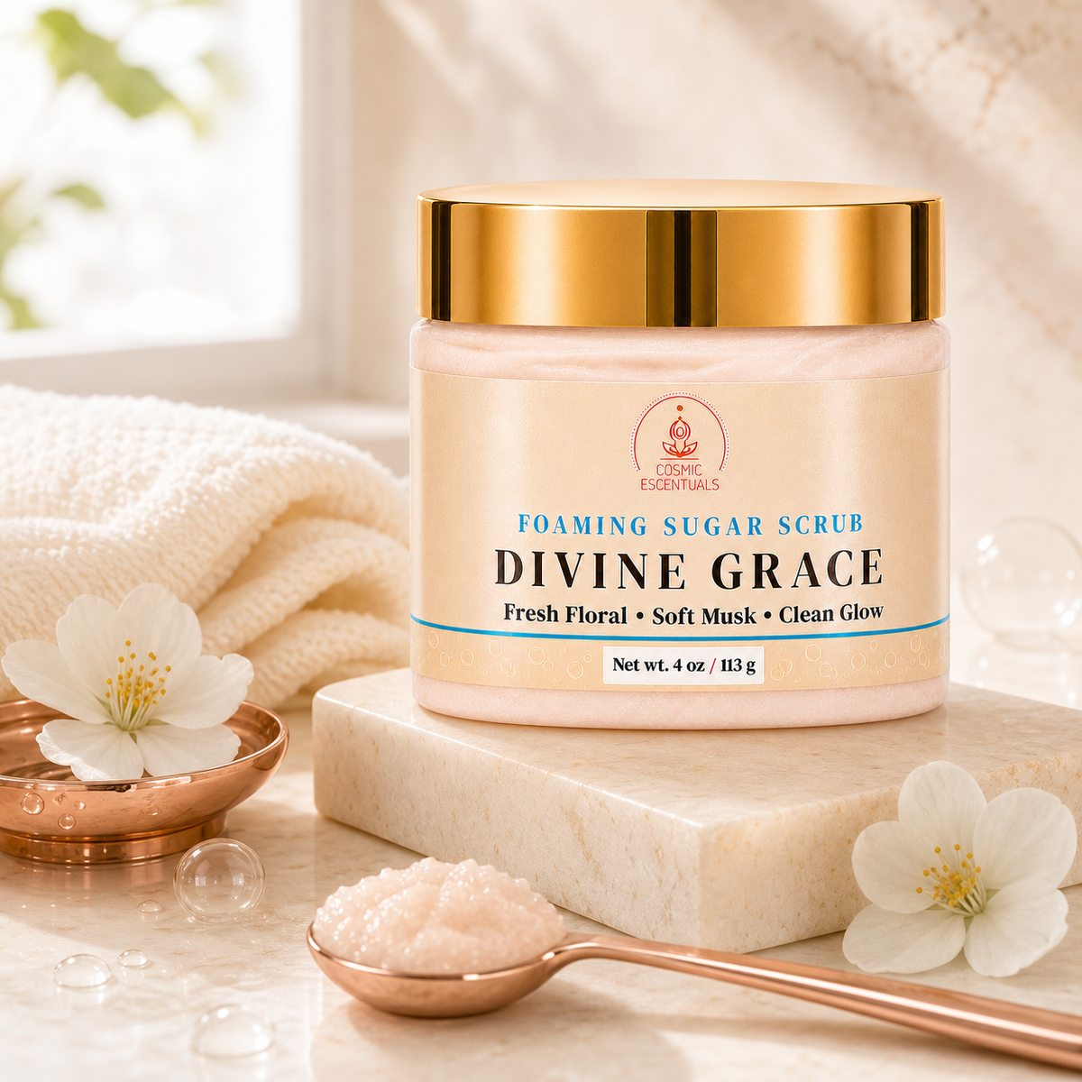 A jar of Cosmic Essentials Divine Grace Foaming Sugar Scrub sits on a white marble block, surrounded by white flowers, a copper spoon with scrub, and soft cream-colored towels in a bright, clean setting.