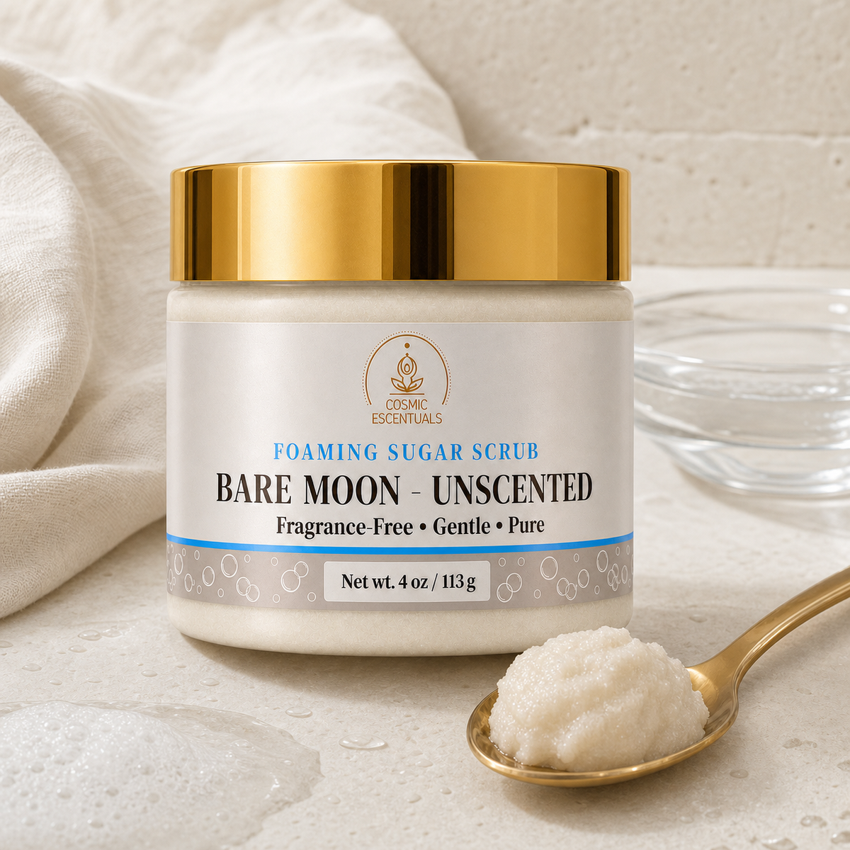 A jar of Cosmic Essentials Bare Moon Unscented Foaming Sugar Scrub, shown on a textured beige stone surface with a gold spoon holding some of the white scrub and a soft beige fabric in the background.