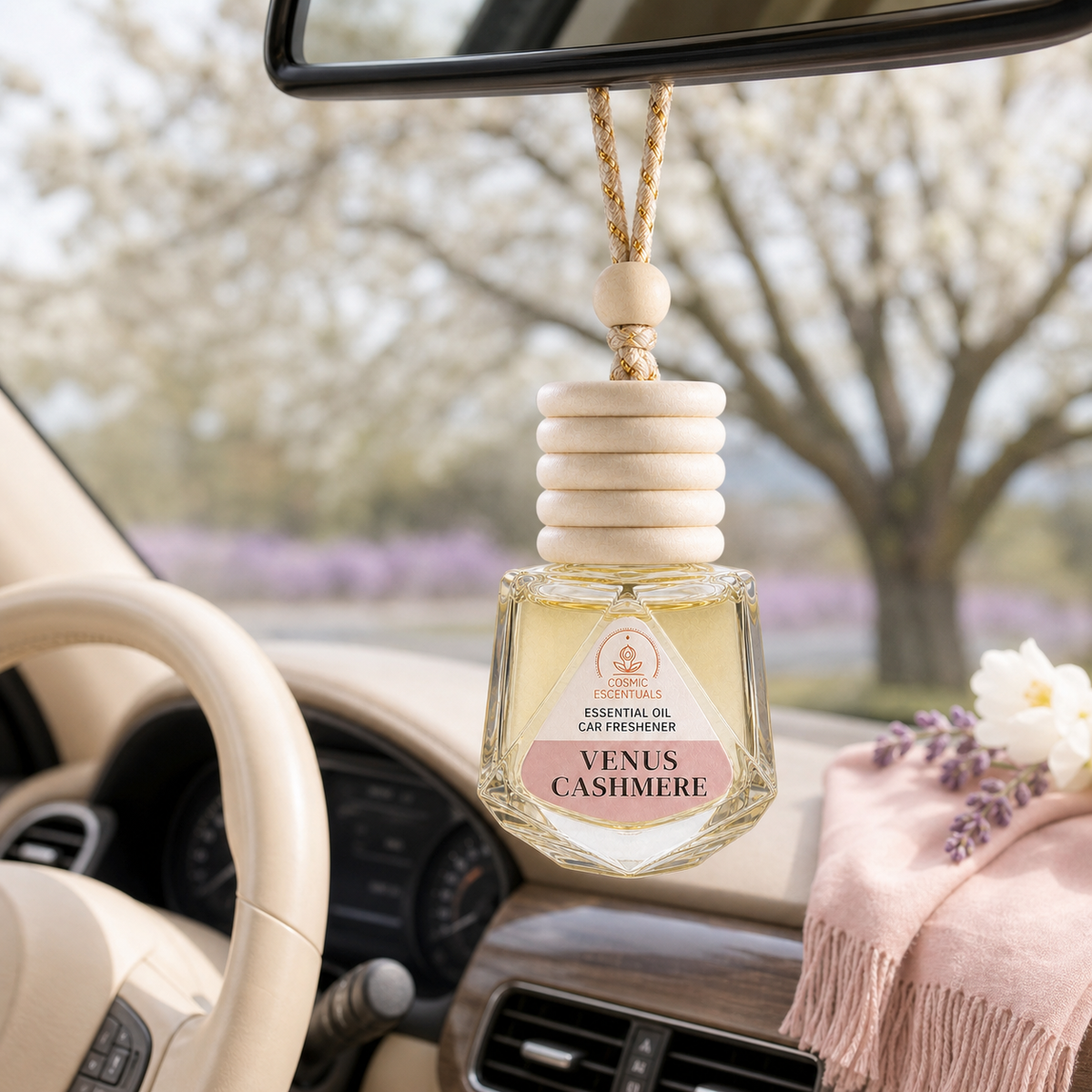 A Venus Cashmere essential oil car freshener bottle with a wooden diffuser top hangs from the rearview mirror inside a car. The bottle is clear glass containing a pale yellow liquid, with a cream-colored label that reads 'Venus Cashmere'.
