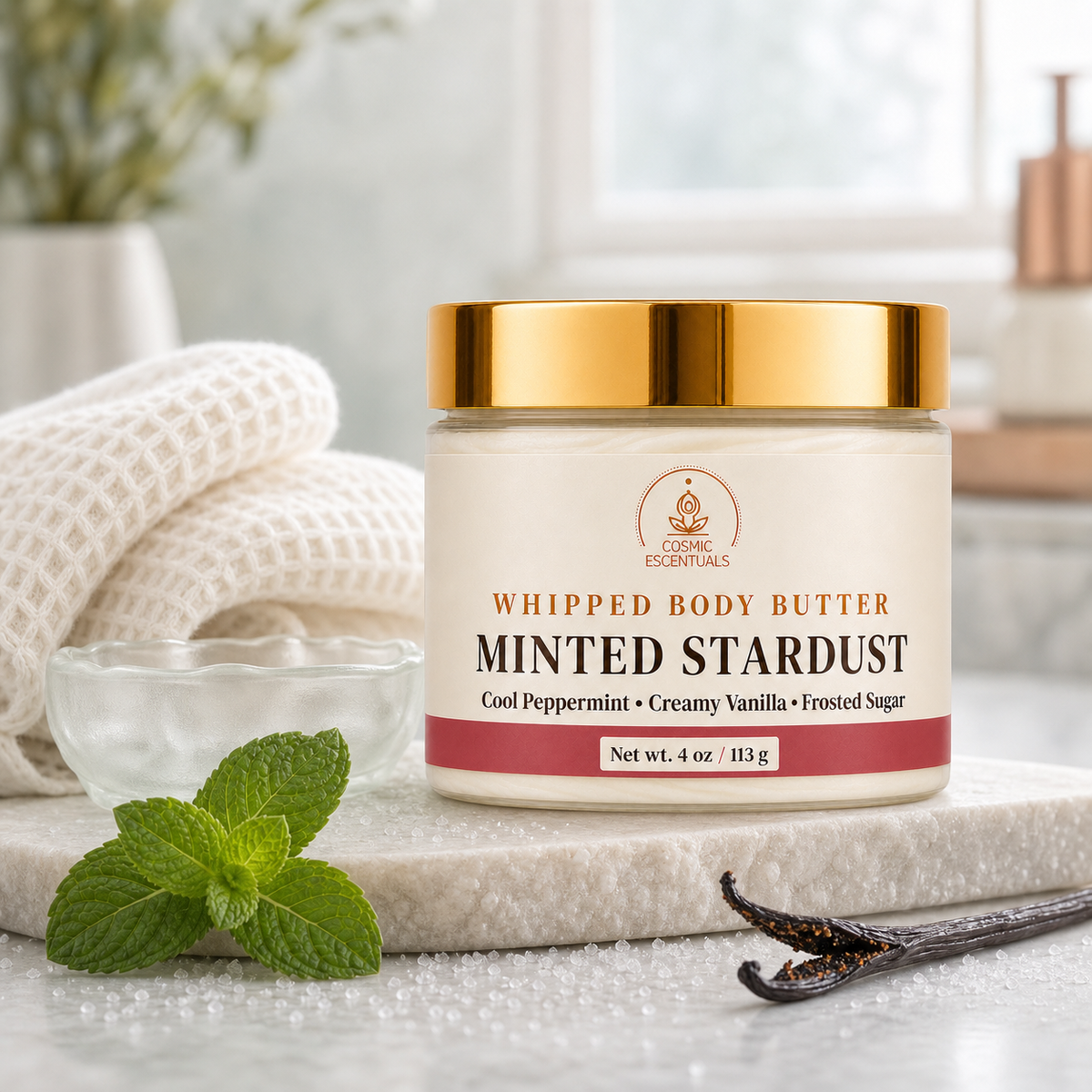 A jar of Cosmic Escentuals Minted Stardust whipped body butter sits on a stone slab with mint leaves, a vanilla bean, and a small glass dish in a bright, softly blurred bathroom setting.