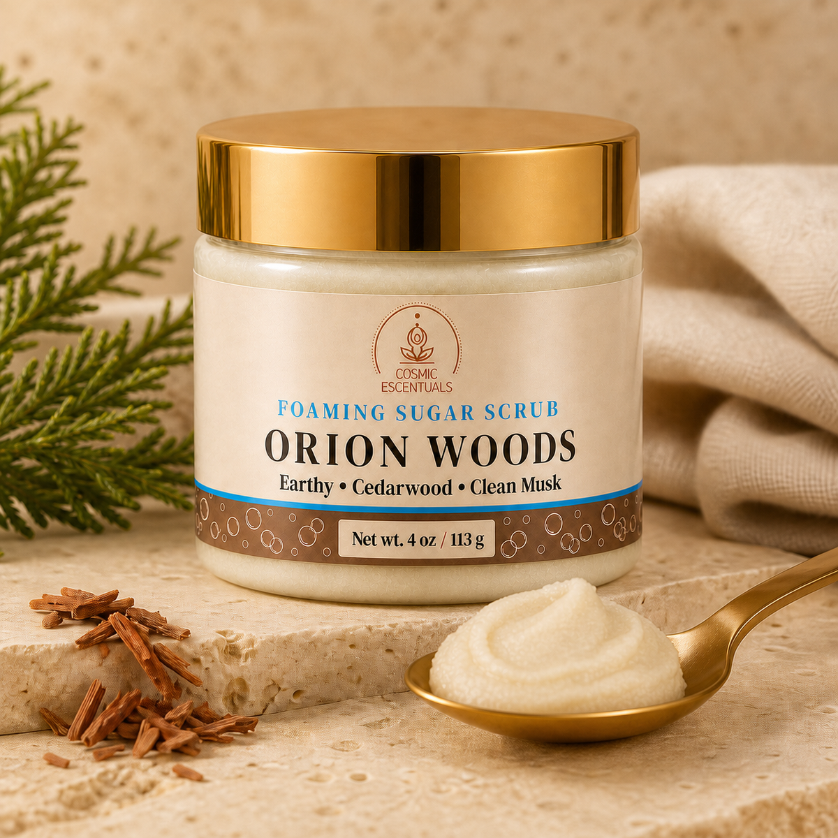 A jar of Organic Essentials Orion Woods Foaming Sugar Scrub sits on a stone slab with a gold spoon of the white scrub in front, surrounded by cinnamon sticks, greenery, and a beige cloth.