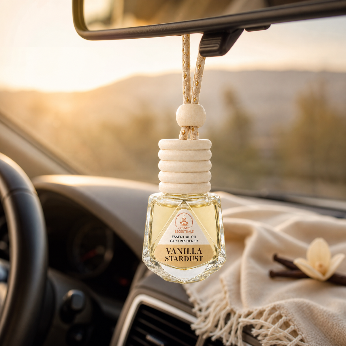 Vanilla Stardust Essential Oil Car Freshener hanging from a rearview mirror at sunset, with a cream scarf draped over the dashboard.