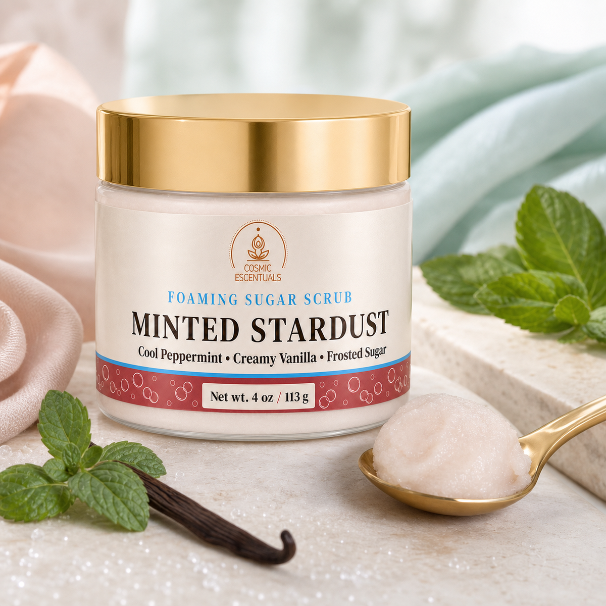 A jar of Cosmic Escentuals Minted Stardust Foaming Sugar Scrub, styled with mint leaves, a vanilla bean, a gold spoon with cream, and soft draped fabrics on a marble surface.
