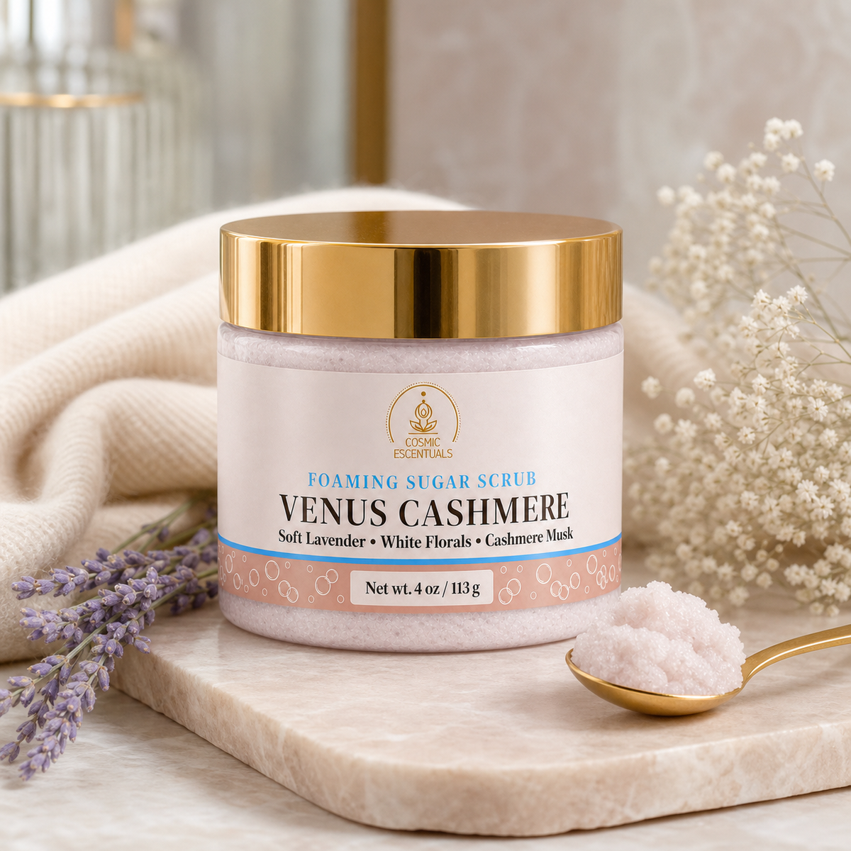 A jar of Cosmic Coordinates Venus Cashmere Foaming Sugar Scrub sits on a marble tray, surrounded by lavender sprigs, a gold spoon with scrub, and soft neutral textiles in the background.