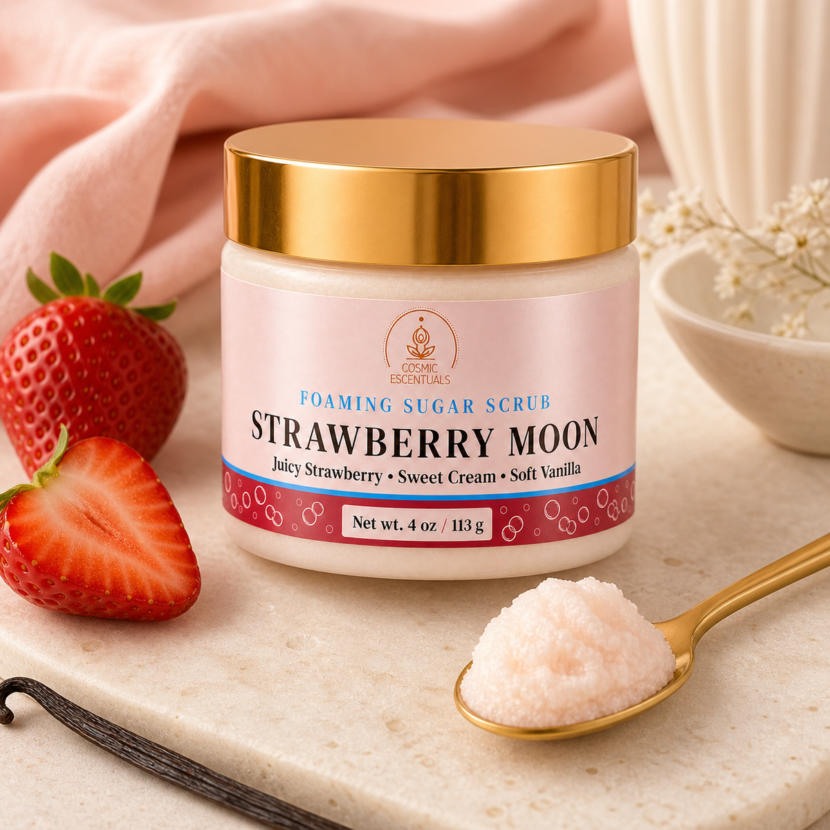 A jar of Cosmic Escentuals Strawberry Moon Foaming Sugar Scrub sits on a marble surface with fresh strawberries, a gold spoon holding some of the scrub, and a pink cloth in the background.