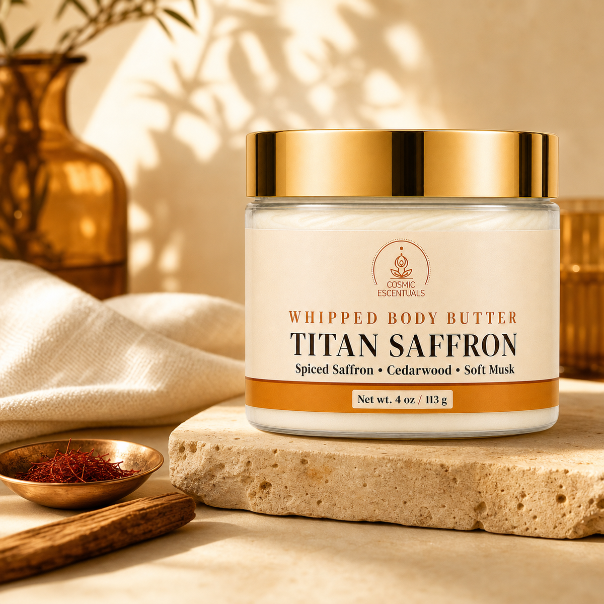 A jar of Cosmic Essentials Titan Saffron Whipped Body Butter sits on a stone slab in a warm, styled setting with a cream cloth, a small copper bowl of saffron threads, and a cinnamon stick. The jar has a white label with gold and brown text, a gold lid, and is set against a warm beige background with soft shadows.