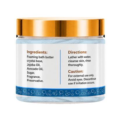 A jar of foaming bath butter with a metallic gold lid and a white label displaying ingredients, directions, and caution details. The jar is filled with light blue bath butter crystals and features a blue patterned band near the bottom.
