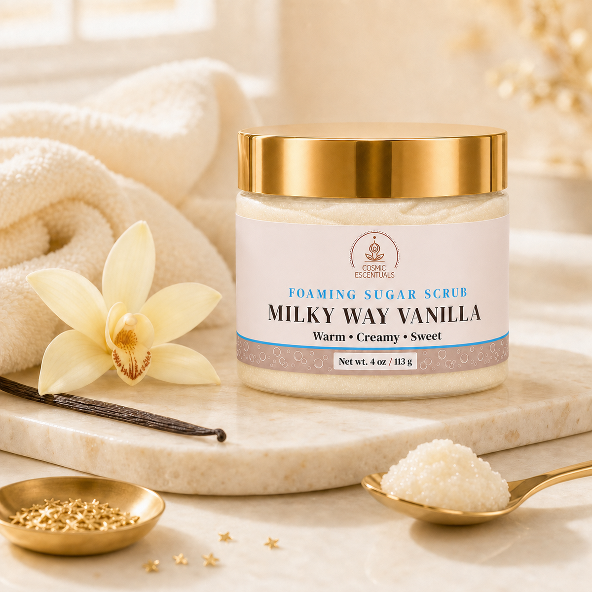 Milky Way Vanilla Foaming Sugar Scrub styled with vanilla beans, a vanilla flower, towels, and gold spa accessories.